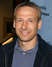 Head and shoulders of a man wearing a open-collar light blue shirt and a navy blue fleece. He has short blonde hair, blue eyes, and stubble.