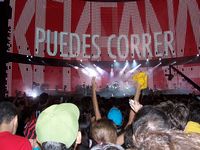 The view of the stage from the audience's perspective, surrounded by fans, with the words "PUEDES CORRER" visible on the video screen.  A camera on the right side shows the concert being filmed.