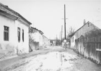 A black-and-white photograph of a street, one-story white houses on either side, a fence on the right in the foreground, the road appeared unpaved, and muddy. There are no people. There is what appears to be a stop sign, a pole with a sign on the top, but no words are visible. There are some tall poles along the street, perhaps telegraph poles.