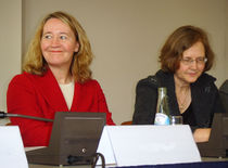 Two women in their fifties sitting at a table with a microphone on it. The woman to the left is dressed in red and is smiling. The woman to the right is wearing glasses and dark clothes.