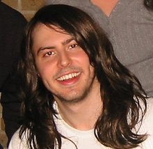 A close-up of a Caucasian adult male with long brown hair smiling.