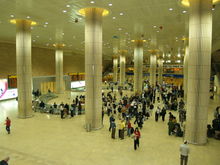 A large hall, with tall round columns; dozens of people standing or walking, around an entrance in the left wall. The area around the entrance is partitioned for the most part from the rest of the hall.