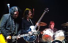 Prominent in the foreground, two guitarists concentrate on their playing, while the drummer, a little behind them to their left, toils away.