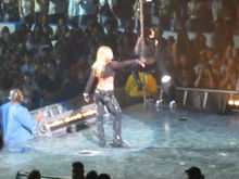 Image of a blond female performer. She is turning her back to the camera, near the end of the stage. She is wearing black pants and a black top with long sleeves, showing her midriff. In front of her, a female dancer is climbing into a pole. A crowd of people clapping, screaming and taking pictures stands can be seen in the audience.