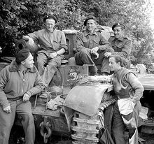 Three soldiers sit on a tank while two more stand next to it.