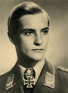 The head and shoulders of a young man, shown in semi-profile. He wears a military uniform with an Iron Cross displayed at the front of his white shirt collar. His hair appears blond and short and combed back, his nose is long and straight, and his facial expression is determined; looking to the left of the camera.