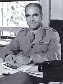 A black and white photograph of a man in formal military uniform sitting at a desk. He has short, thinning hair and large bushy eyebrows.