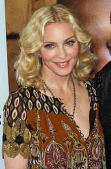 Upper body of a middle-aged blond woman. Her hair is parted in the middle and falls in waves to her shoulder. She is wearing a loose dress with black and brown prints on it. A locket is hung around her neck, coming up to her breasts. She is looking to the right and is smiling.