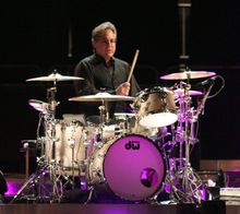 Middle-aged man with slightly graying dark hair and glasses wearing black shirt sits behind a silver-colored, purple-lit drum kit with his right hand and stick about to hit a snare drum or cymbal.