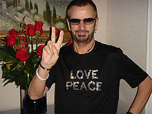 A man in his sixties wearing sunglasses and a black T-shirt bearing the sequined words "LOVE" and "PEACE". Only his torso is visible, but his left hand appears to be on his hip. His right hand forms a V-shaped peace symbol in reference to his quote "Peace and Love".