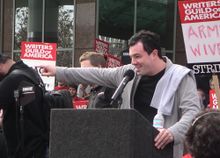 A man wearing a gray jacket and black shirt, with black hair and light skin, points with his right hand with his arm fully extended.