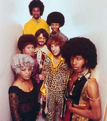 Seven young adults in garish clothes and hair. The most prominent is a black man in a vest with chains; he wears an enormous afro with sideburns, and looks with narrowed eyes and closed mouth at the camera.  A black woman is in a gray wig and black dress. A white man with red hair wears a leopard print shirt and pants. There are two other black men, also in afros, another white man, with a short beard and glasses, and another black woman.