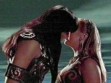 On the left is a tall black haired woman, dressed in leather. She is bending down to kiss a young, slightly shorter , blonde haired woman.