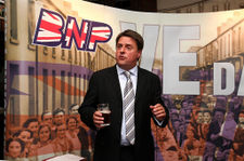 A three-quarter length shot of a suited man standing with a glass of beer in his right hand, in front of a large hoarding displaying a montage of photographs, and the BNP logo