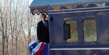 Obama is standing aboard the back platform of a train and looking to the side of the train.  There is a red, white and blue banner hanging over the rear railing.