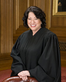 A formal pose from waist up of a smiling, light-medium-skinned woman with somewhat curly black hair of almost shoulder length, dressed in a black judicial robe and standing behind a chair with hands folded on the chair, in formal-looking room with medium brown wood paneling and a bit of painting on a wall.