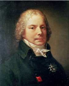 Portrait of a man, showing his head and shoulders. He is wearing a black jacket with a high-necked collar and a white shirt tied in a bow. His head is adorned by a curly blonde wig.