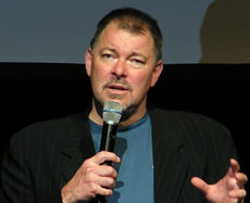 Bearded man in a black jacket gesturing while talking into a microphone.