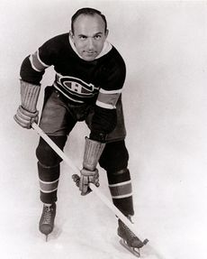 An ice hockey player leans forward with his stick. He has a round face with one eyebrow raised and a bare head with a receding hairline. He wears skates, gauntlets, and a sweater with a stylized "C" around a smaller "H".