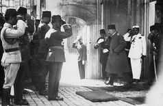 A dozen soldiers salute a man with his head slightly downward as he walks toward a small archway in a wall