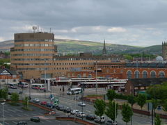 A bus station in front of office buildings. There are hills in the background and a road in the foreground.