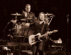 Middle-aged man with glasses wearing a dark shirt sits behind a drum kit on a riser with his right hand and stick about to hit a snare drum; he his looking directly at another middle-aged man in front of and slightly to the left of him, dressed in dark clothes with an electric guitar strapped to him, left hand on the frets, right hand flying away after playing a chord, eyes shut in an expression of conveying musical intensity of some kind