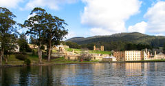 A calm body of water is in the foreground. The shoreline is about 200 metres away. To the left, close to the shore, are three tall gum trees; behind them on an incline are ruins, including walls and watchtowers of light-coloured stone and brick, what appear to be the foundations of walls, and grassed areas. To the right lie the outer walls of a large rectangular four-storey building dotted with regularly spaced windows. Forested land rises gently to a peak several kilometres back from the shore.