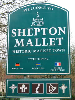 Green signs with white writing and symbols saying Welcome to Shepton Mallet, Historic Market Town, Twin Towns Misburg, Bollnas, Oissel sur Seine