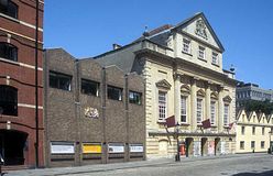 An imposing eighteenth century building with three entrance archways, large first floor windows and an ornate peaked gable end above. On the left a twentieth century grey brick building with a gilded crest and on the right a cream coloured building with four pitched roofs. In front a cobbled street.