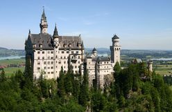 A castle of fairy-tale appearance sitting high on a ridge above a wooded landscape. The walls are of pale stone, the roofs are of steep pitch and there are a number of small towers and turrets.