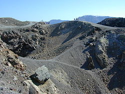 Small figures of people can be seen at the top  looking into  craters strewn with grey rocks.