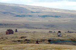 The ruins of a city as viewed from Armenia. A grassy plain is in the foreground with around 10 buildings visible. A hill which is also covered in grass rises out of the plain in the background.