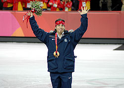 A man wearing a gold medal smiles with his arms raised above his head holding a flower bouquet in his left hand while wearing a dark blue tracksuit and a red bandanna on his head. There is a portion of the ice-rink in the background.