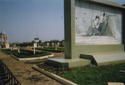 A park scene. A white structure on the left is topped by a blue dome. There is a concrete path and some grass. On the right, there is a large structure bearing a black-and-white drawing of the man and boy from the scenes described above, crouching, the man waving with his left hand, the boy holding onto the man's t-shirt.