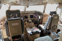 A flight deck, from behind the two pilots' seats. A center console lies in between the seats, in front is an instrument panel with several displays, and light enters through the forward windows.