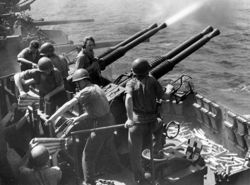 The gun crew operating these guns; four men handle ammunition while another yells over the blast from the guns. Spent shell casings litter the deck below.