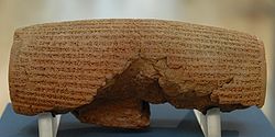 Front view of a barrel-shaped clay cylinder resting on a stand. The cylinder is covered with lines of cuneiform text