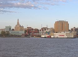 A row of tall buildings in the middle, with a river on the bottom and a blue cloudy sky on top