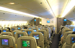 Airliner cabin. Rows of seats arranged between two aisles. Each seatback has a monitor; light shines from the sidewalls and overhead bins.