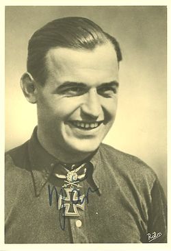 The head and shoulders of a young man, shown in semi-profile. He wears a shirt with an Iron Cross displayed at the front of his shirt collar. His hair is dark and short, his nose is long and straight, and his facial expression is showing a broad smile; gazing at a point to the right of the camera.