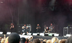 Five men play instruments onstage; one sings, one plays a drum set, one plays bass guitar, and two play electric guitar.  An audience watches the band play.