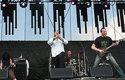 Four men are shown on stage, from left to right, one man plays bass guitar, while another man sings into a microphone, the man shown in the back plays on a large drum kit while the man on the far right plays guitar.