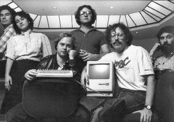 A black and white photograph of five men and one woman, some with long hair and thick glasses, posing alongside a Macintosh.