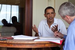 Obama, wearing a white shirt, is moving his hands while talking to a man in a blue shirt, who sits across him.