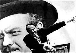 A black-and-white image of a man wearing a suit standing at a podium. His right arm is resting on the podium and is left is pointing up to his left. A large headshot of the same man is visible in the background.