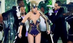 A woman wearing a metallic leotard and helmet is seen walking with a pair of crutches. Behind her, a number of dancers are visible, who are wearing tuxedos with white gloves, and making different gestures behind the woman.