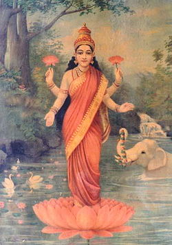 A woman wrapped in a red sari, with long hair and four hands stands on a red lotus and holds two red lotuses in her two upper hands.