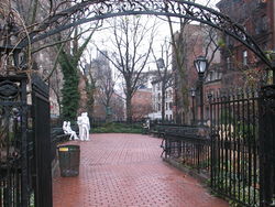 A color photograph of Christopher Park in winter, showing the wrought iron entrance arch in the foreground and the brick pavement surrounded by five and six story brick buildings; in the center background are four white statue figures: two males standing, one with his hand on the other's shoulder, and two females seated on a park bench, one woman with her hand touching the other's thigh. All are dressed in jeans and loose clothing