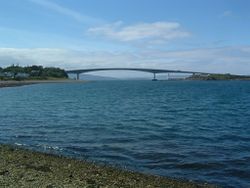  A body of blue water is spanned by a concave bridge of modern design in the middle distance. A small lighthouse can be seen beyond the bridge under its span.