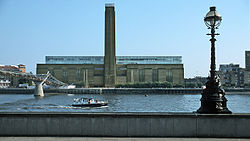 A large oblong brick building with square chimney stack in centre of front face. It stands on the far side of a river, with a curving white foot bridge on the left.